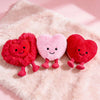 Three small, soft plush hearts in red and pink are displayed on a fluffy pink background.