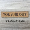 Un patch tissé brun avec le texte YOU ARE OUT gravé, montré sur un fond de bois gris.