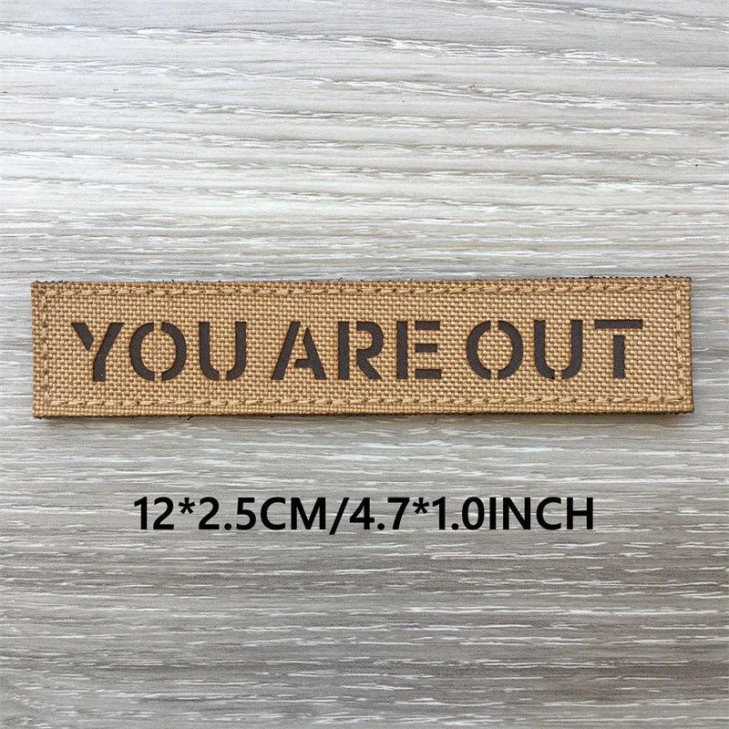 Un patch tissé brun avec le texte YOU ARE OUT gravé, montré sur un fond de bois gris.