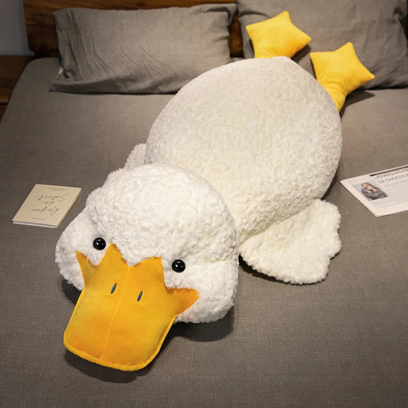 A white plush duck pillow with yellow beak and yellow accents resting on a gray fabric surface.