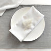 A white linen dinner napkin is placed on a white plate on a gray linen cloth.