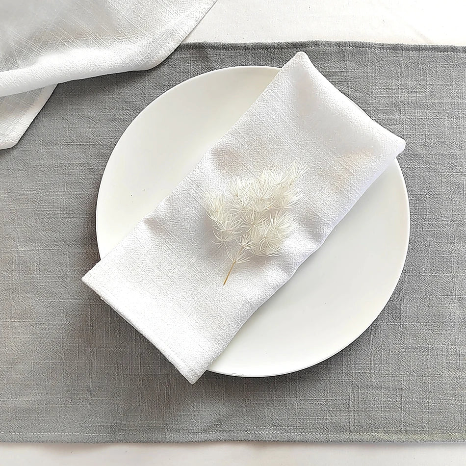 A white linen dinner napkin is placed on a white plate on a gray linen cloth.