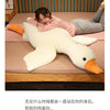A person is cuddling a large white plush goose pillow with orange feet.