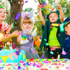 Two children are celebrating outdoors with colorful confetti, including a girl in a green cape and a girl with a pink bow.