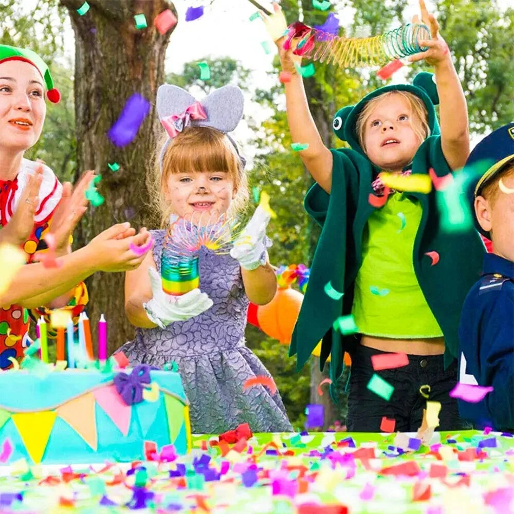 Two children are celebrating outdoors with colorful confetti, including a girl in a green cape and a girl with a pink bow.