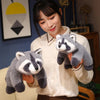 A woman is holding a gray and white raccoon plush toy against a light-colored sweater.