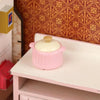 A pink miniature copper soup pot with a lid displayed on a white shelf against a patterned background.
