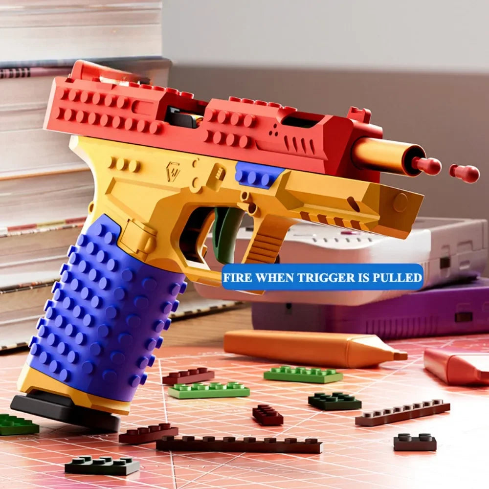 A yellow and red building block toy gun with a blue firing mechanism is displayed on a surface of small building blocks.