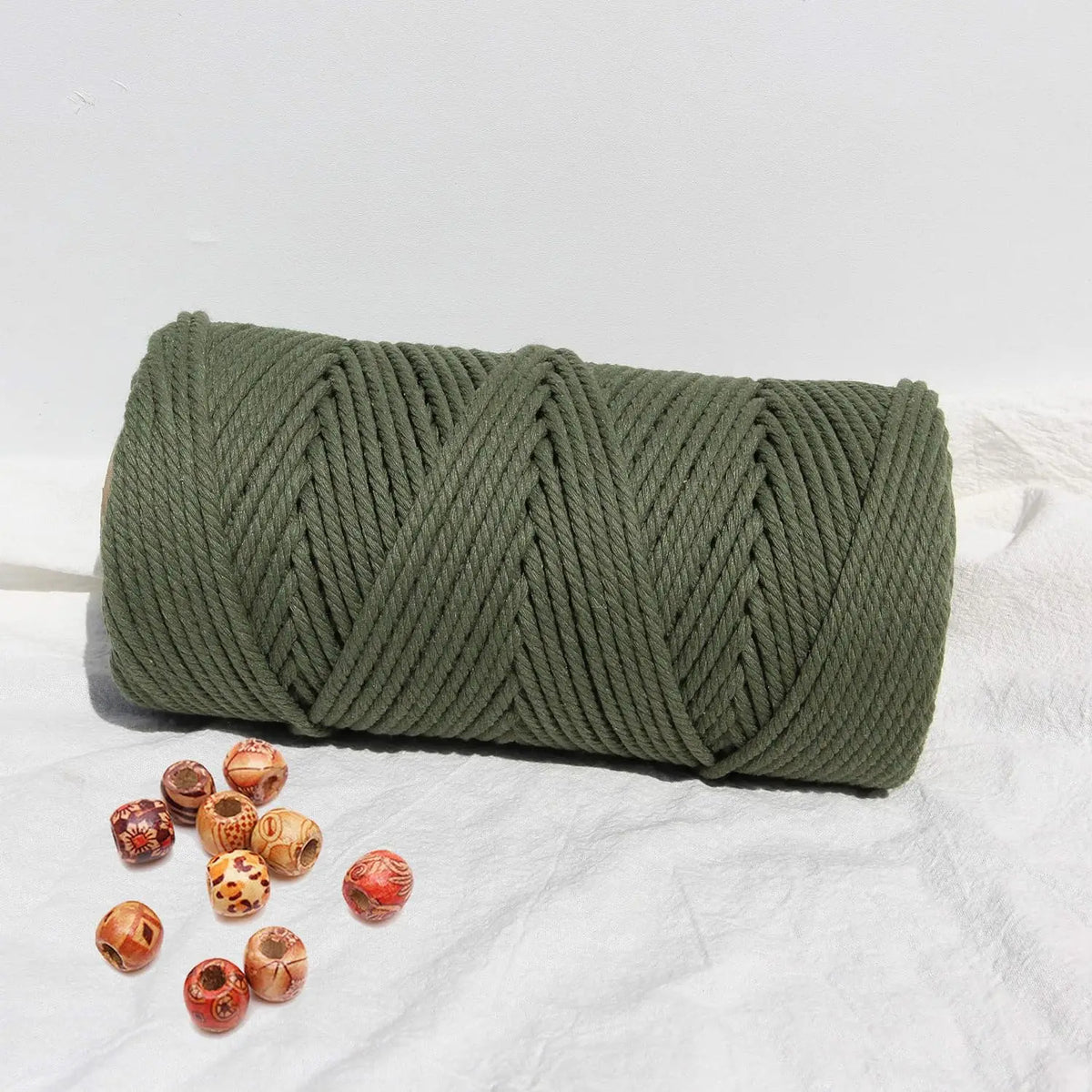 A coil of natural cotton macrame rope in olive green resting on a white surface with small wooden beads.