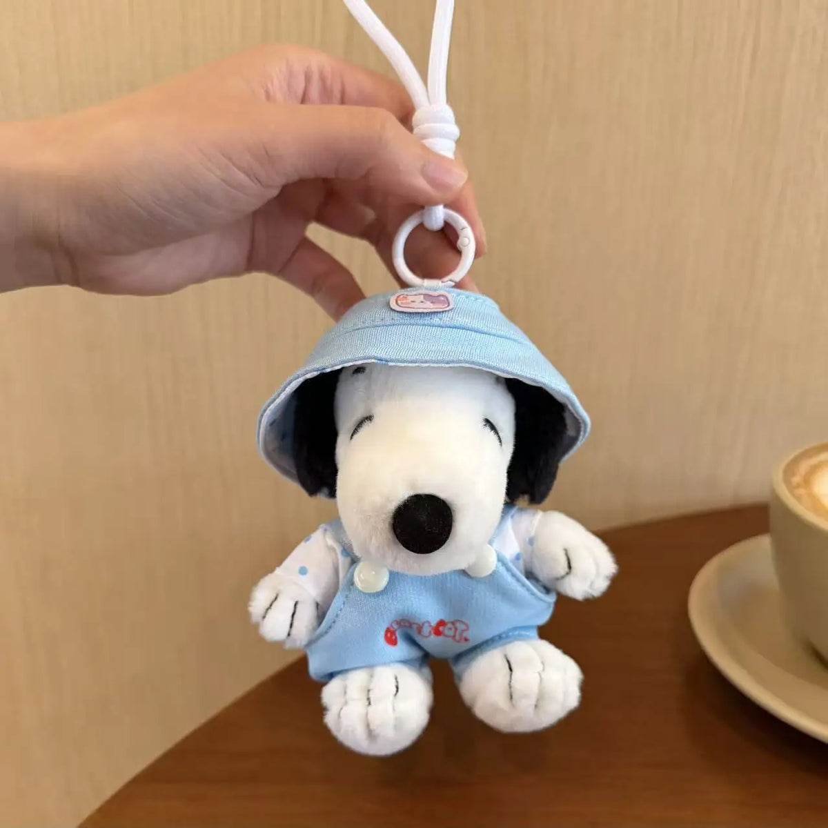 A light blue Snoopy plush charm wearing a hat is being held by a hand, ready to be attached to a bag.