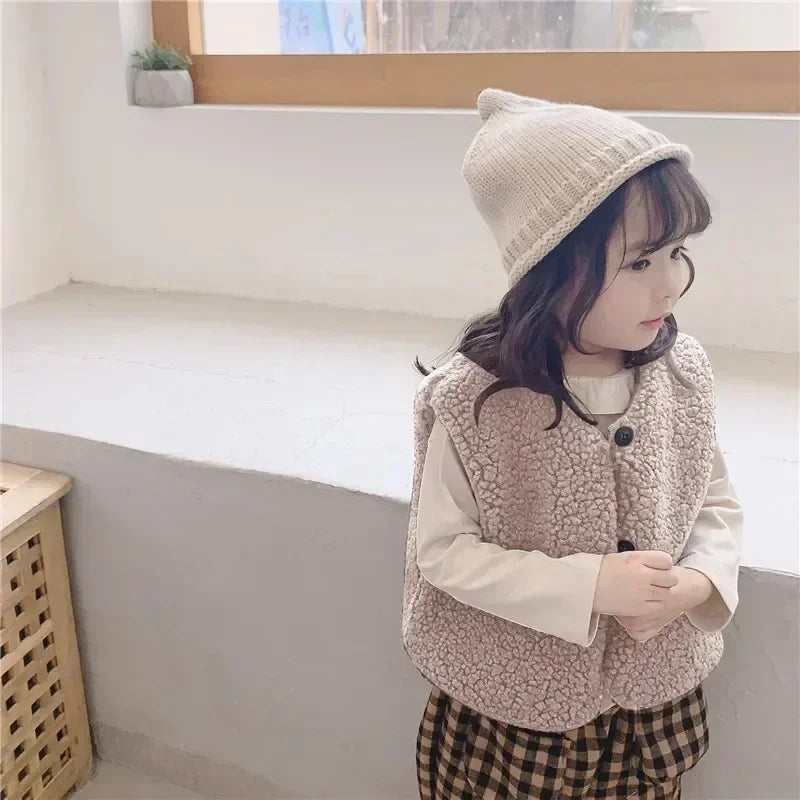 A young girl wearing a beige knitted hat and a light brown fleece vest over a white shirt and plaid skirt.