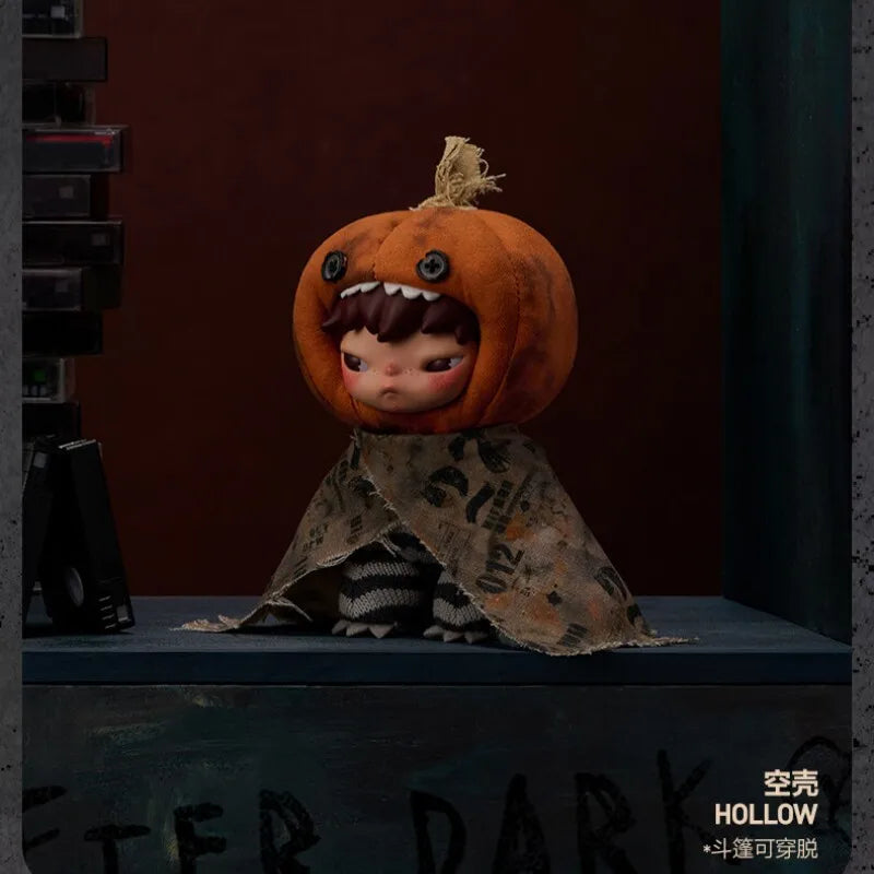 An orange plush doll with a stitched mouth and hair, wearing a patterned fabric drape, displayed on a dark surface.