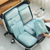 A light blue travel packing cubes and storage bag set are shown open on a white rolling suitcase.
