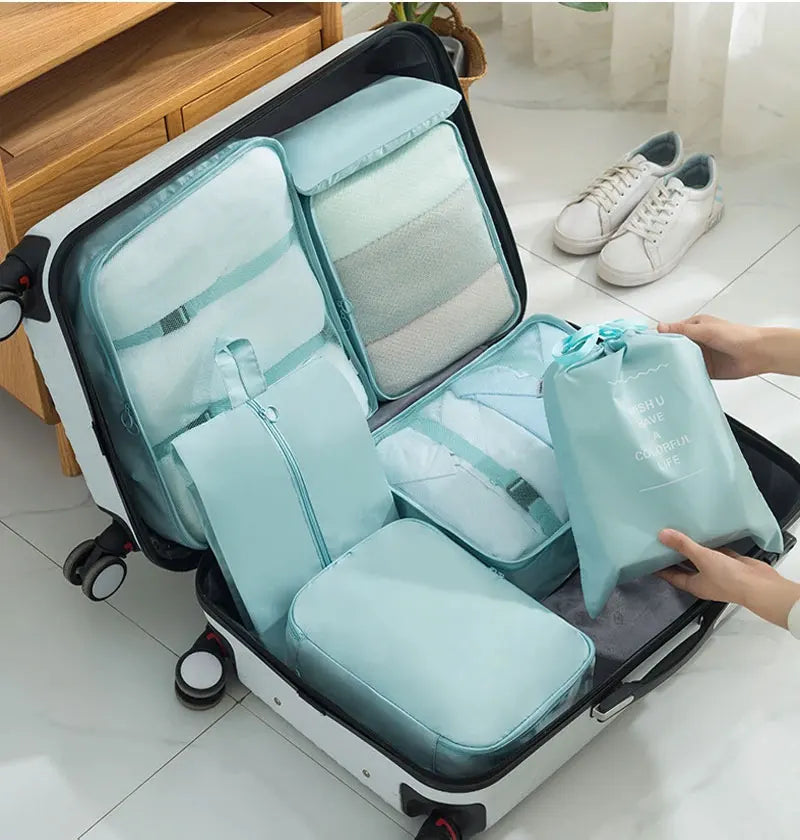 A light blue travel packing cubes and storage bag set are shown open on a white rolling suitcase.