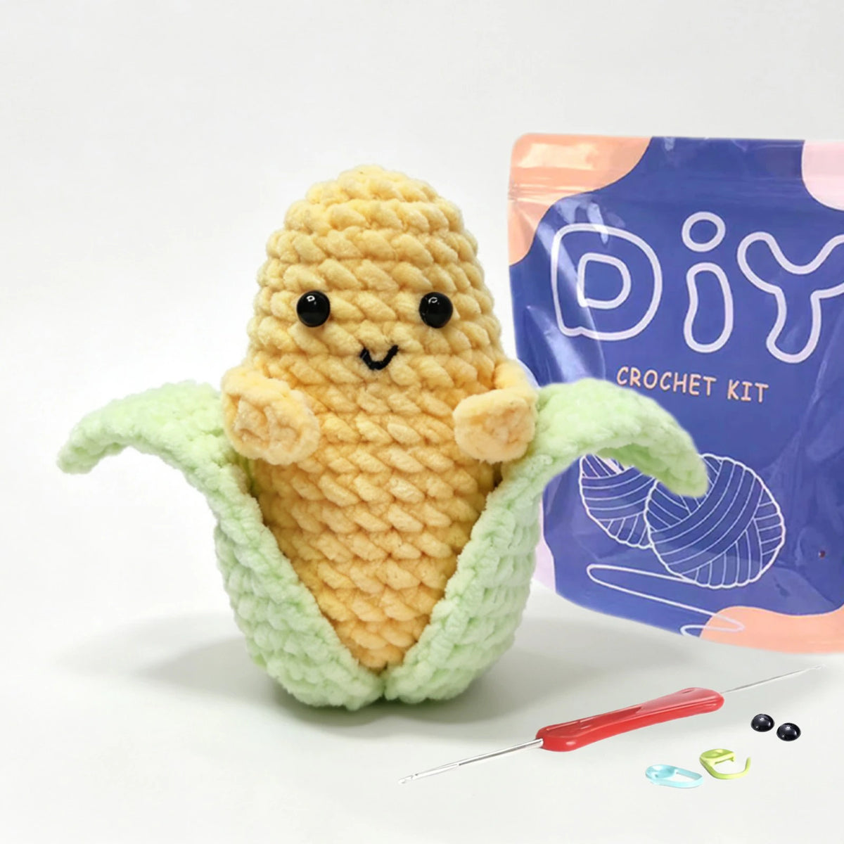 A yellow crocheted egg shape with green leaves is displayed next to its purple packaging and a crochet needle.