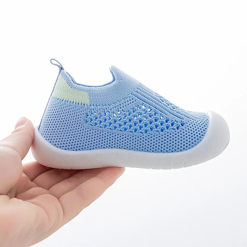 A hand holds a light blue breathable mesh slip-on sneaker for toddlers.