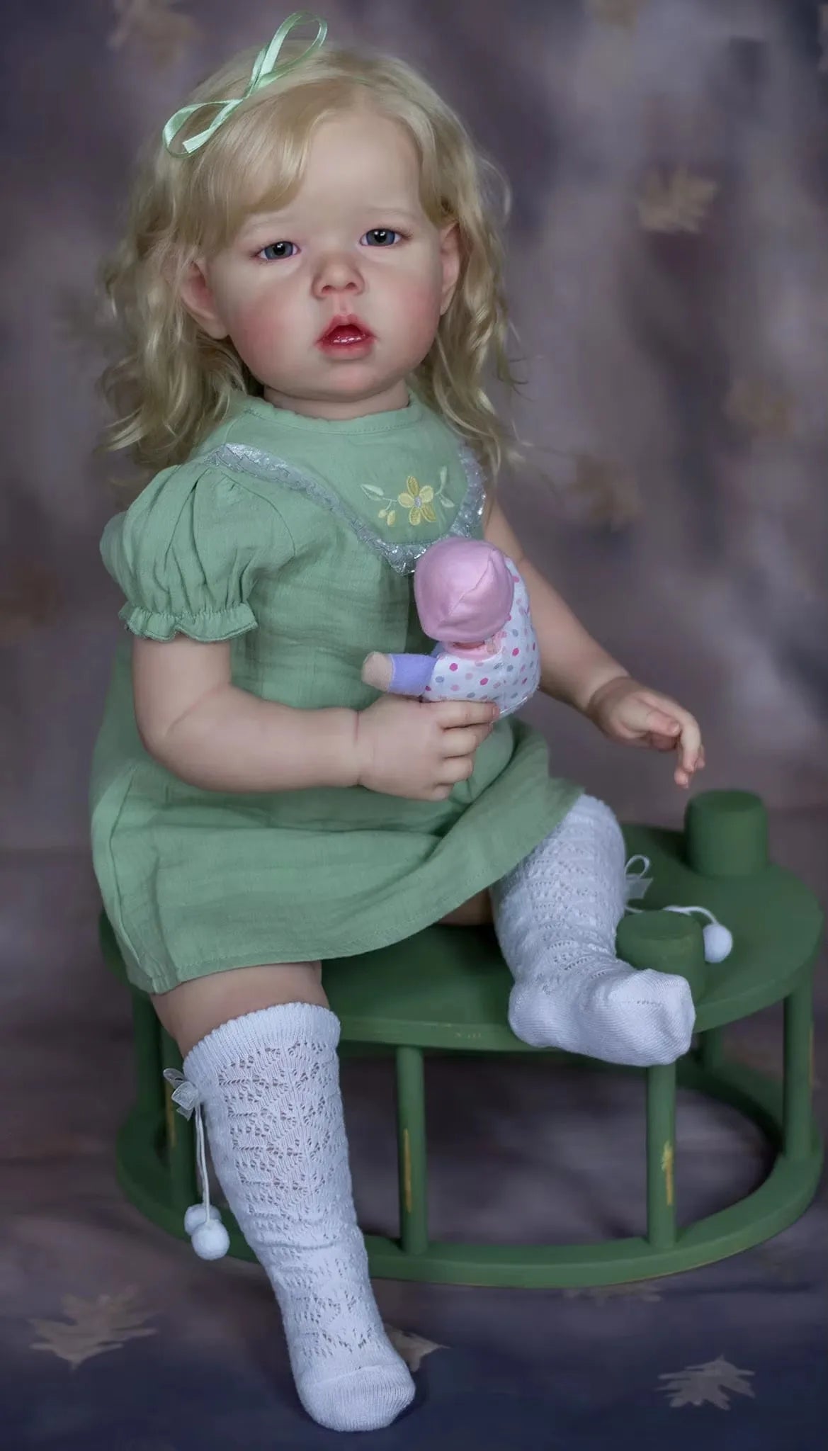 A realistic reborn baby doll with soft cloth body wearing a green dress and white socks.