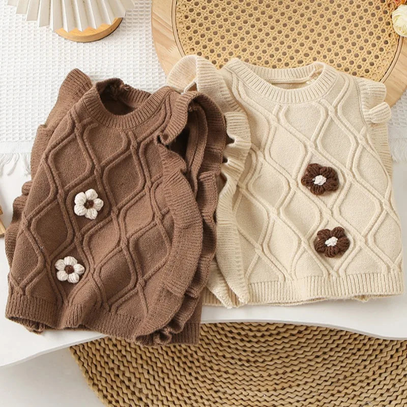 A brown knit sweater vest for girls with a ruffled edge and flower-shaped details, displayed on a woven mat.