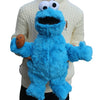 A bright blue Cookie Monster plush toy with large eyes and an open mouth is displayed against a white sweater.