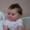 A close-up photo of a lifelike reborn baby doll named Maddie with soft brown hair and pink clothing.