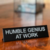 A black desk sign with white text that reads Humble Genius at Work, resting on a wooden surface.