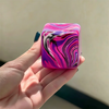 A hand holds a vibrant purple and pink marbled cube toy with swirling black lines.