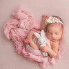 A newborn baby is swaddled in a pink knit wrap with a pearl headband, resting on a soft pink background.