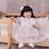 A baby doll with soft cloth body wearing a floral dress and hair accessories, posed in a wooden crib.