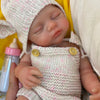 A newborn silicone reborn doll wearing a knitted outfit and hat, held in a yellow and white setting.