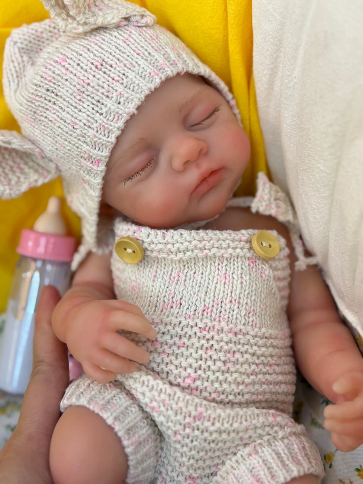 A newborn silicone reborn doll wearing a knitted outfit and hat, held in a yellow and white setting.
