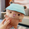 A hand is holding a soft, brown panda bear plush toy wearing a light blue hat.