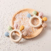 Holzplatte mit Silikon-Fingerfreund-Ring und bunten Silikon-Zahn-Attachments sowie klaren Zuckerkristallen.