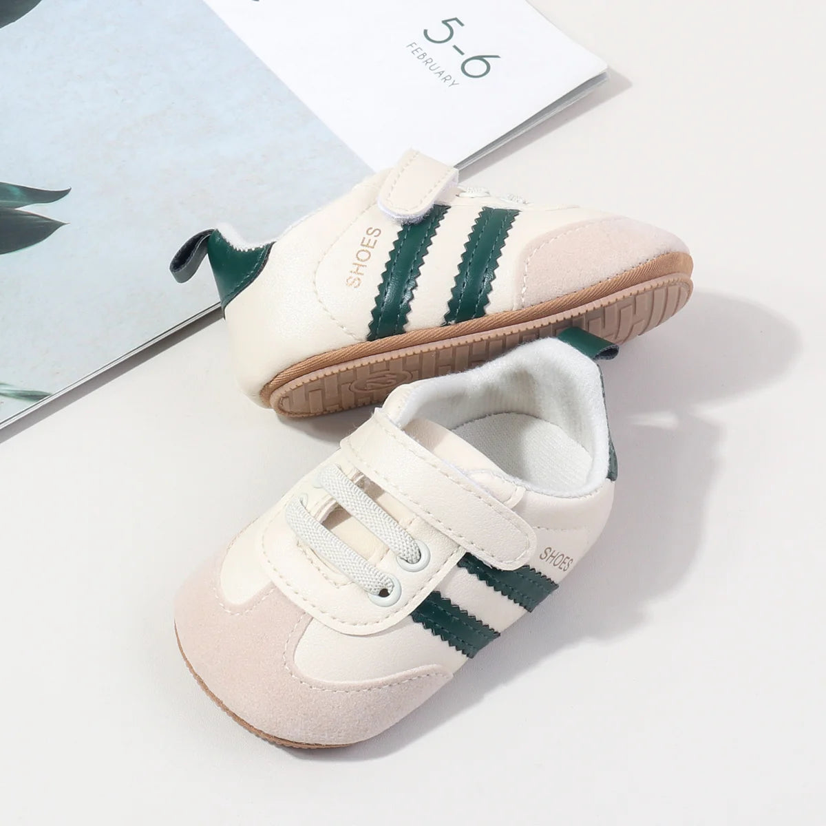 A pair of light pink baby first walkers shoes with green stripes and soft sole, shown next to a card.