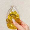 A hand holds a clear glass bottle filled with yellow liquid and several yellow squishy stress balls labeled Chiboki.