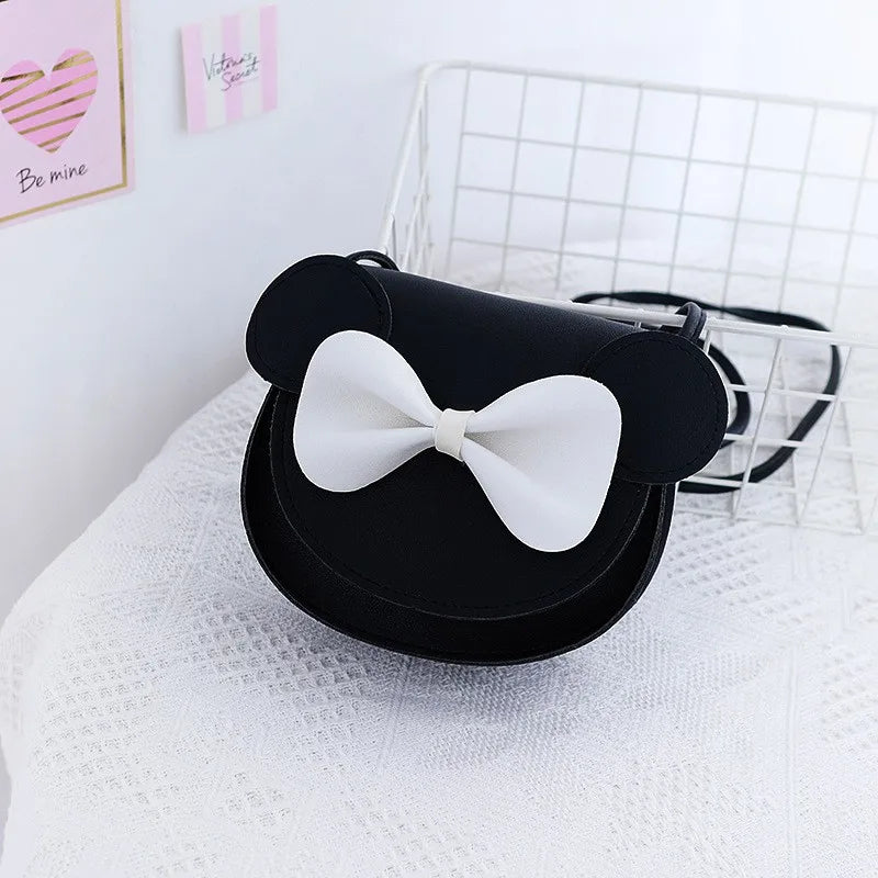 Black kids' hard shell crossbody bag with mouse ears and a white bow resting on a white textured surface.