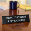 A black desk sign with white text reads OOOOH THIS NEEDS A SPREADSHEET on a wooden surface.