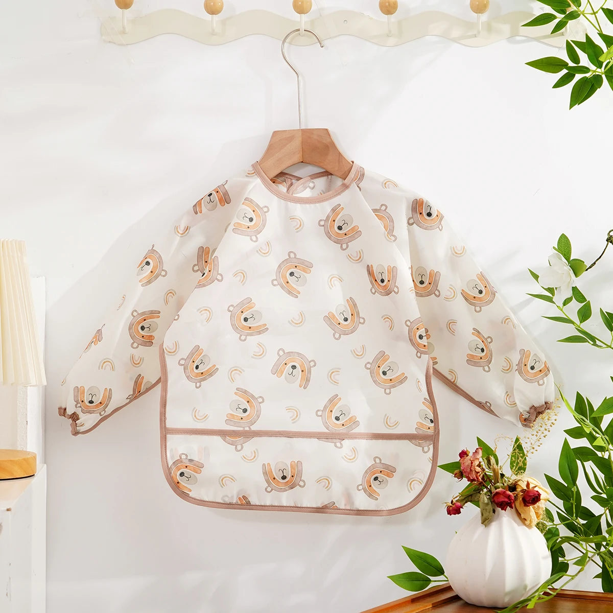 A white waterproof baby bib with long sleeves featuring a pattern of cute bear faces, hanging on a wooden hanger.