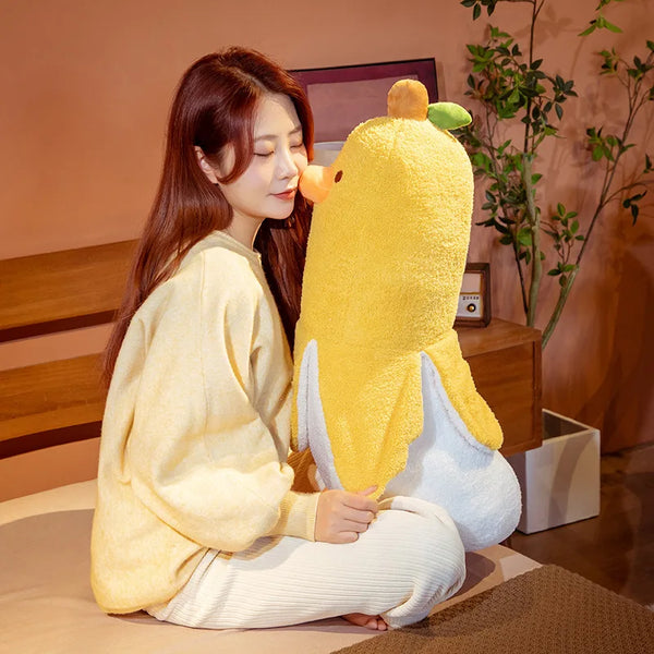 A woman is sitting next to a large yellow banana duck plush pillow.