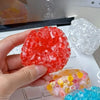 A hand holds a bright red squishy fidget ball next to a clear white one and a multi-colored ball.