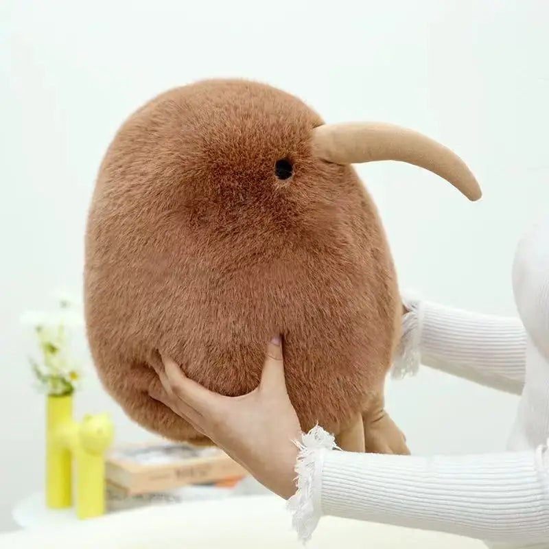 A hand is holding a large, brown kiwi bird plush toy against a white background.