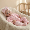 A lifelike soft body baby doll with pink hair bow and pink outfit resting in a textured white basket.