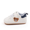 A pair of white baby sneakers featuring a cute brown bear design on the toe and a navy blue lining.