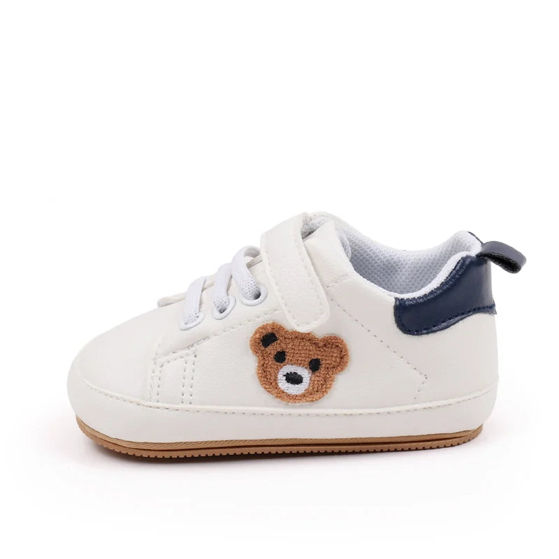 A pair of white baby sneakers featuring a cute brown bear design on the toe and a navy blue lining.