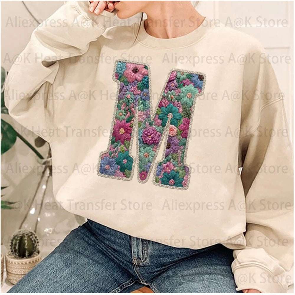 A light beige sweatshirt featuring a large embroidered floral letter M on the chest.