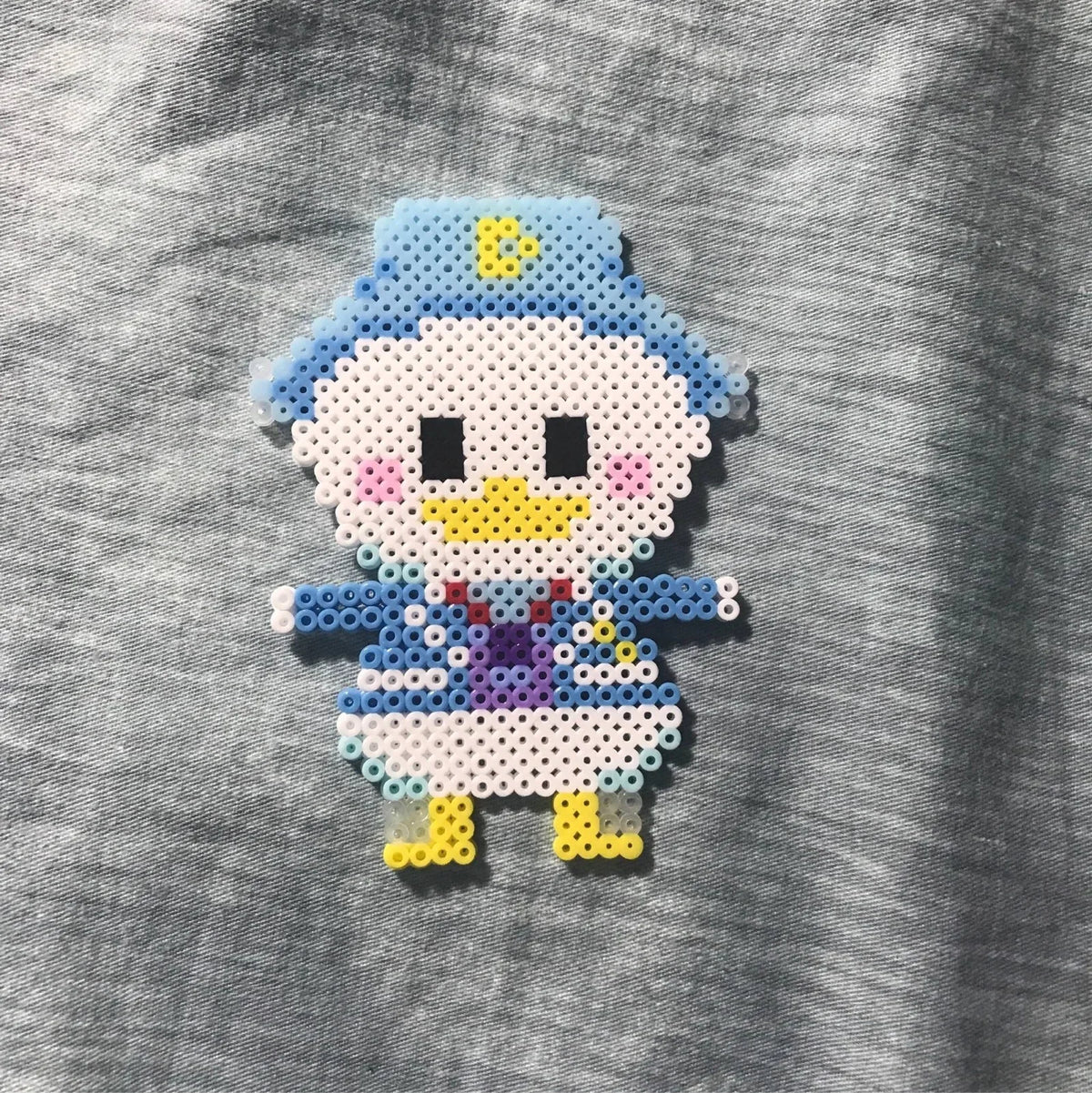 A white and blue fuse bead figure resembling a bird is crafted using small plastic beads on a gray fabric background.