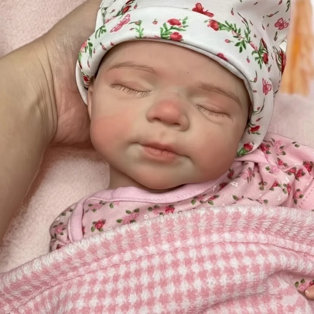 A close-up photo of a lifelike silicone reborn baby doll wearing a floral hat and pink clothing.