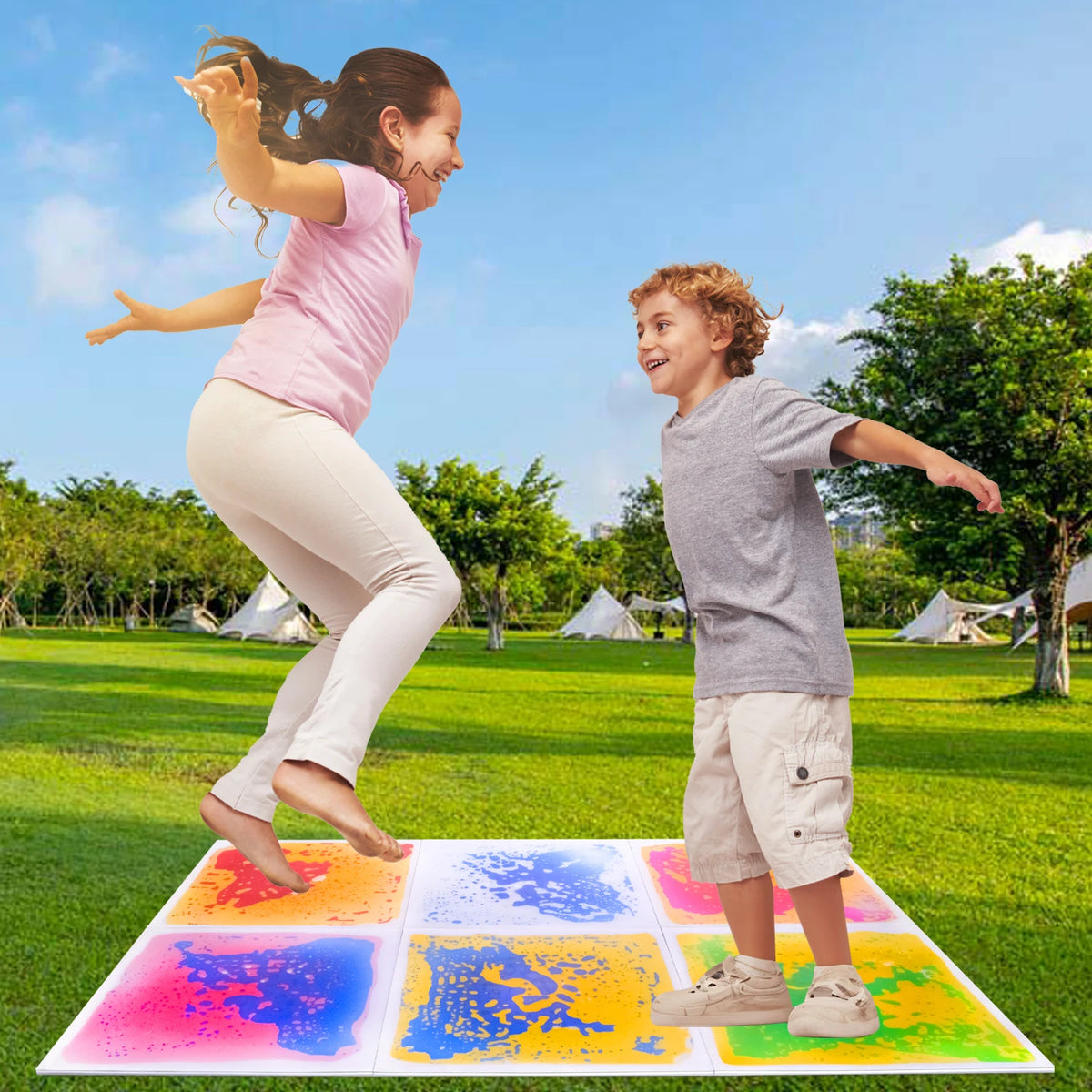 Two children play on a colorful liquid sensory play mat outdoors in a grassy field.