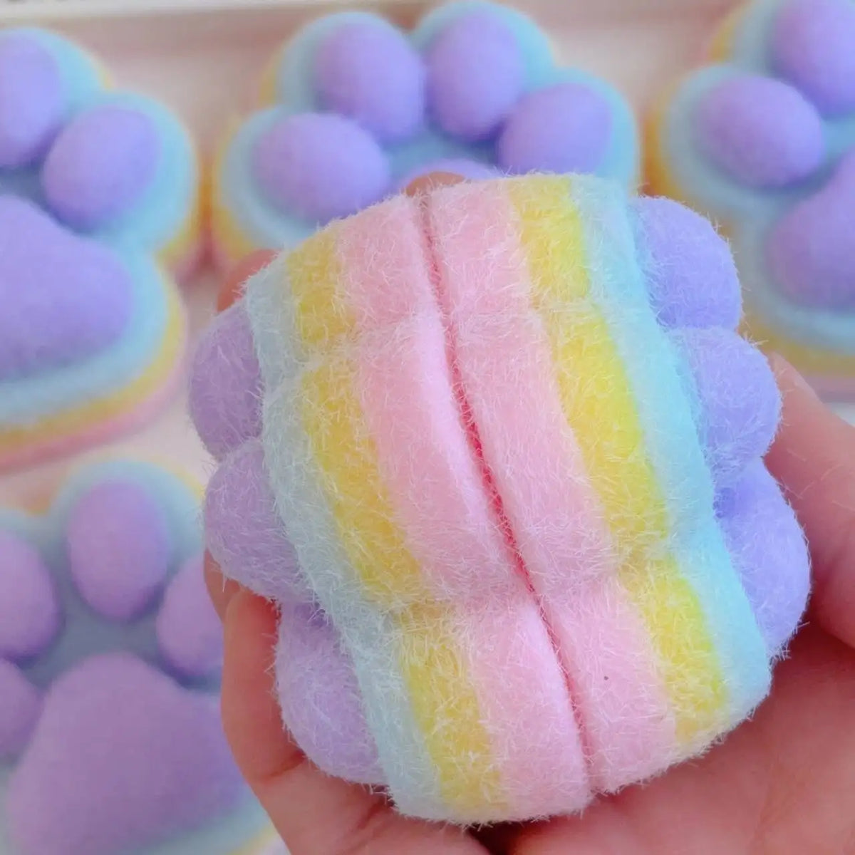 A hand holds a pink and yellow striped squishy toy shaped like a cat paw, surrounded by other purple and green mochi toys.