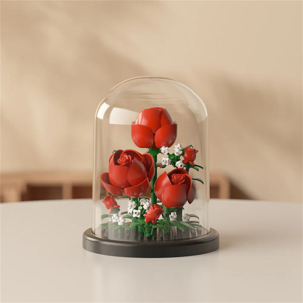 A clear glass cloche displays a miniature bouquet of red roses and white filler flowers.