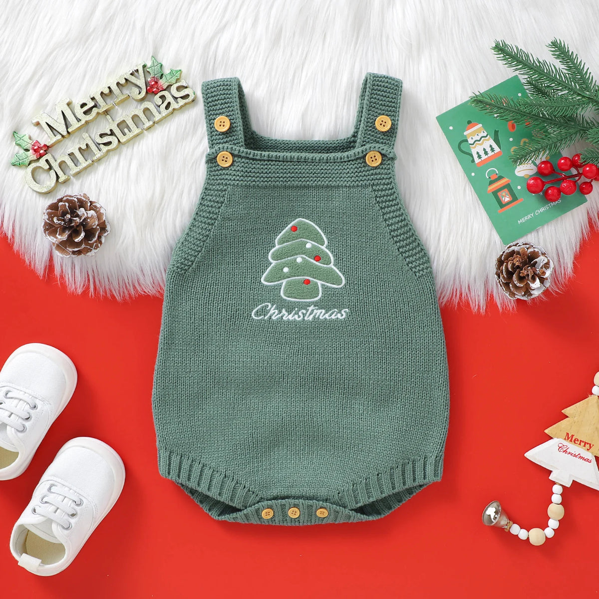 A green knit baby romper with Christmas embroidery lies on a red background surrounded by white boots and pine greenery.